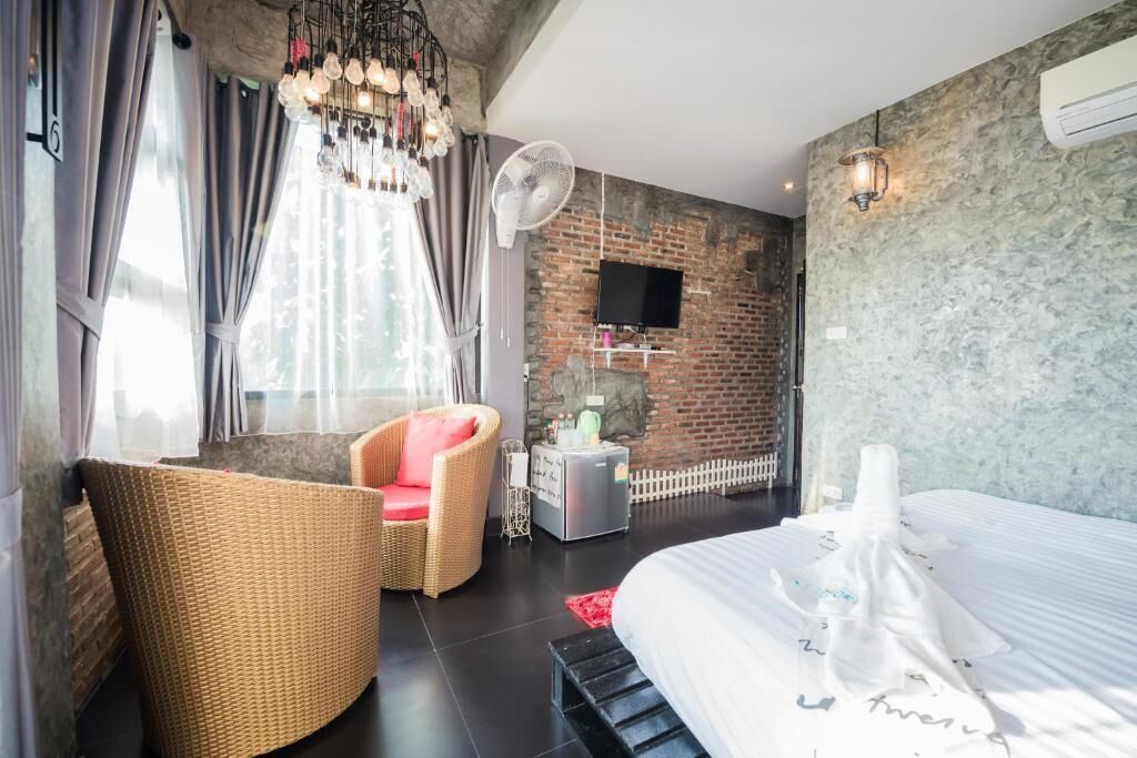 A modern hotel room with a brick wall, a large bed, a wicker chair, a small table with a fan, and a TV mounted on the wall.