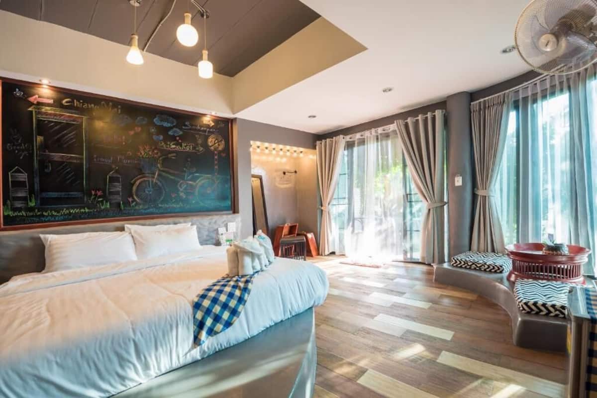 A hotel room with a large bed, a seating area with cushions, a chalkboard wall with drawings, and large windows with curtains.