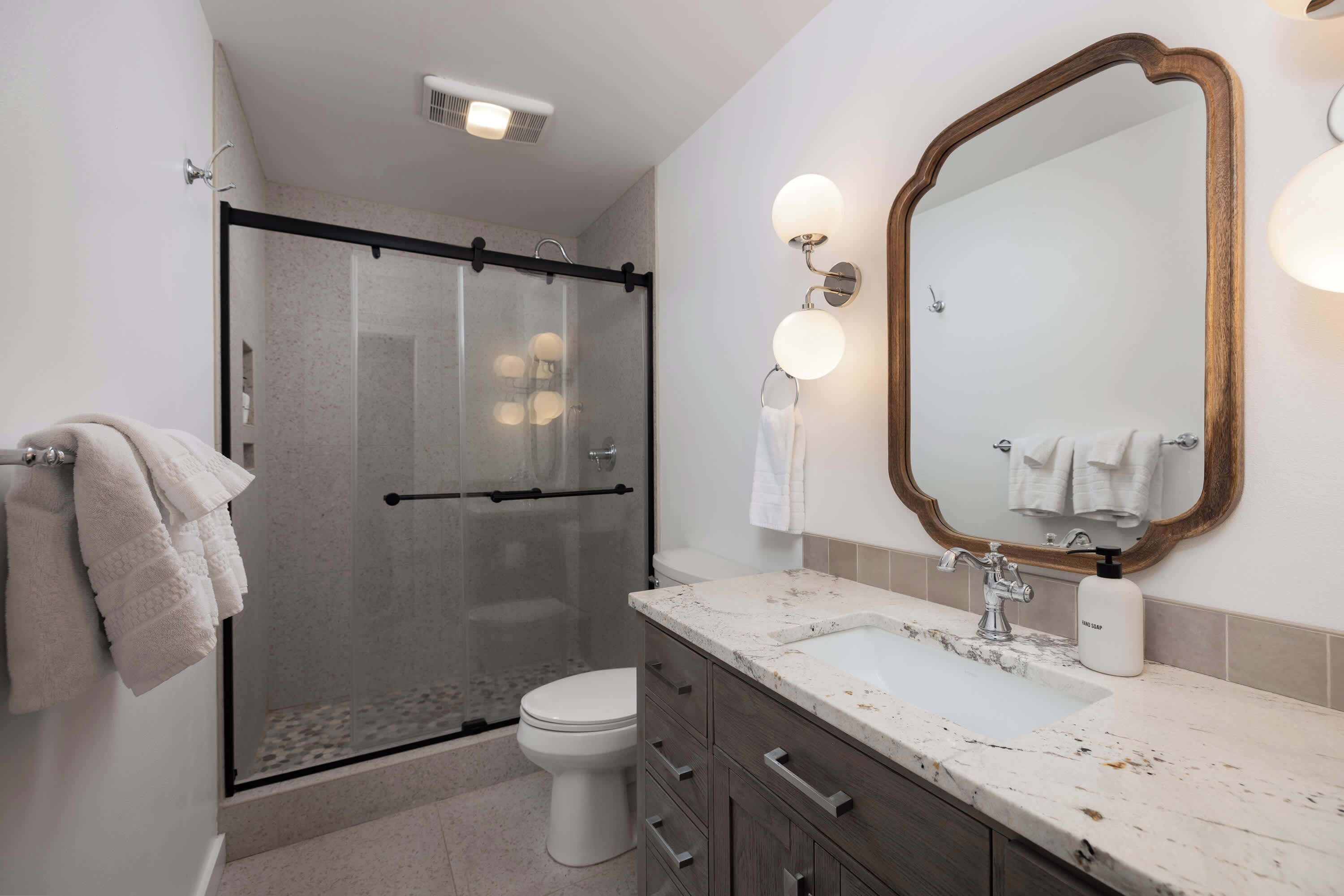 A modern bathroom with a glass-enclosed shower, a white toilet, a marble sink, and a large mirror with wall sconces.