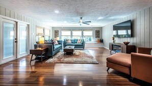 Ein geräumiges Wohnzimmer mit Deckenventilator, großen Fenstern und einem an der Wand montierten Flachbildfernseher.