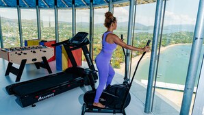 A woman exercising on a cardio machine with a view of a beach and cityscape through large windows. - Résidence La Canopée (Nosy Be)