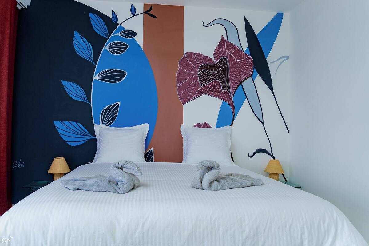 A hotel room with a bed, two pillows, a red curtain, two lamps, and a wall with a floral and abstract design.
