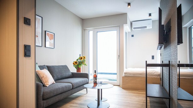 Una sala de estar moderna con un sofá gris, una mesita de centro redonda y una pequeña mesa auxiliar. Hay un ventanal con vistas al exterior, y un televisor de pantalla plana montado en la pared.