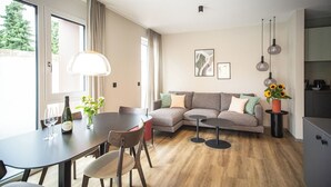 Una sala de estar moderna con comedor, sofá, mesa de centro y mesa de comedor con sillas. Hay dos lámparas colgantes, un jarrón con flores y un cuadro en la pared.