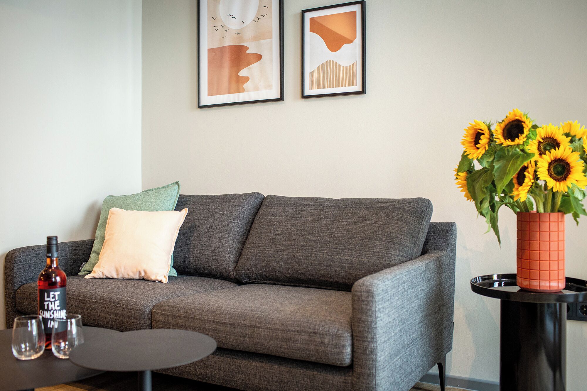 Ein Wohnzimmer mit grauem Sofa, einem kleinen runden Tisch, einer Weinflasche, zwei Gläsern und einer Vase mit Sonnenblumen.