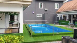 Uma piscina com um deck ladrilhado, cercada por um gramado e um edifício com fachada cinza e várias janelas.
