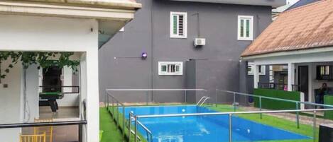 A swimming pool with a tiled deck, surrounded by a grassy area and a building with a grey facade and multiple windows.