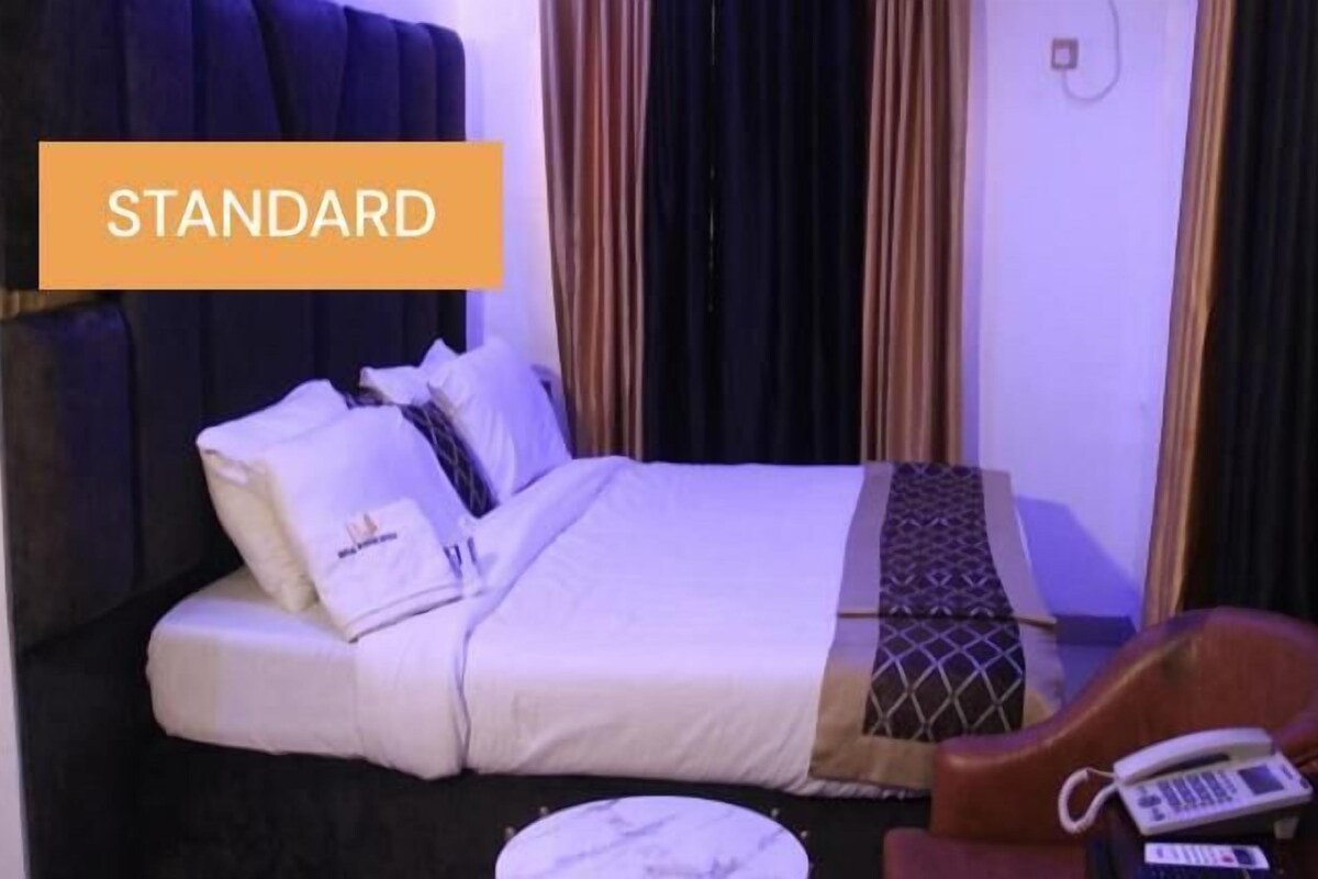 Una habitación de hotel estándar con cama, almohadas, una colcha, una silla y una mesita de noche con teléfono.