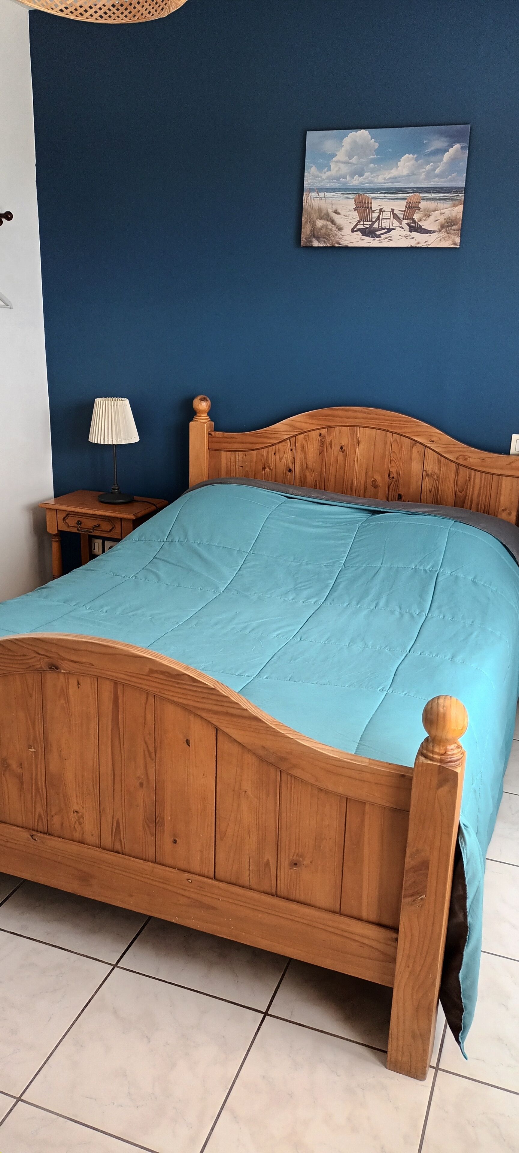 A wooden bed with a blue bedspread in a room with a blue wall and a painting of a beach scene.