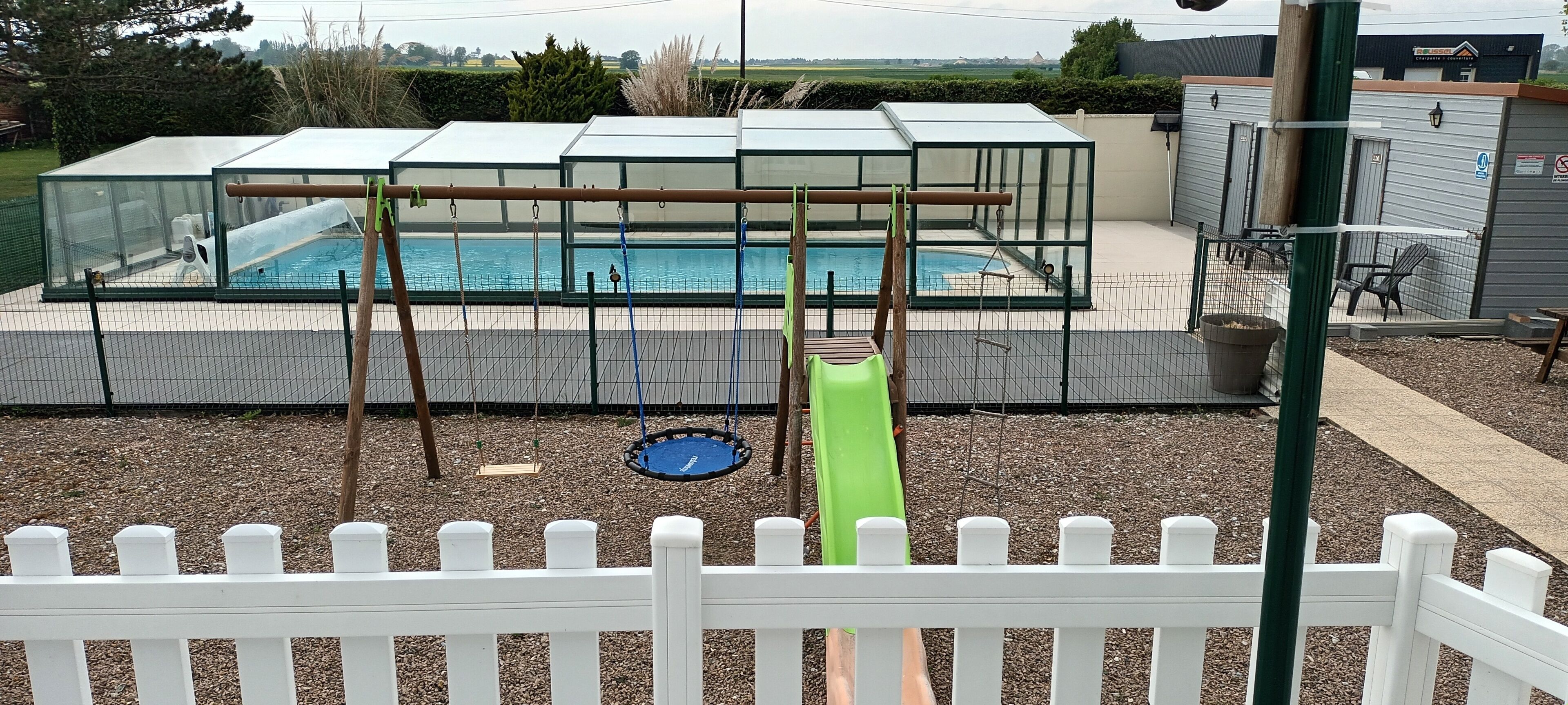 Un espace piscine doté d’un toboggan et d’un trampoline, le tout entouré d’une clôture blanche.