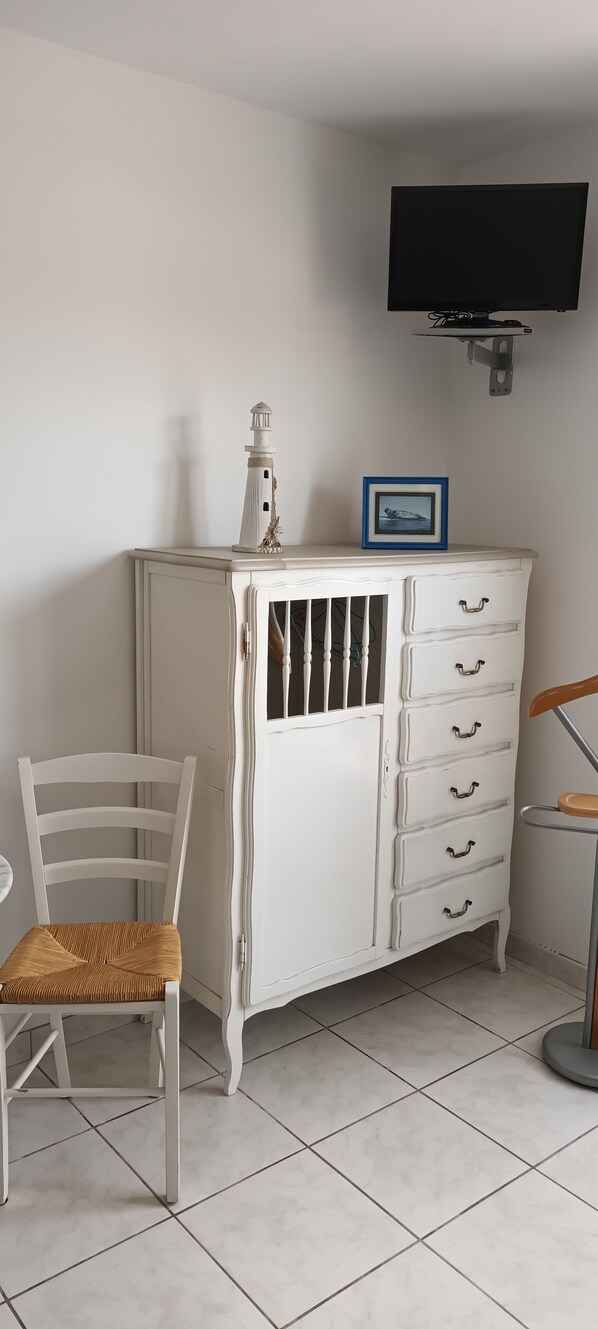 A white cabinet with drawers and a small shelf, a wicker chair, a wooden chair, and a television mounted on the wall. - Apartment with pool (Le Crotoy)
