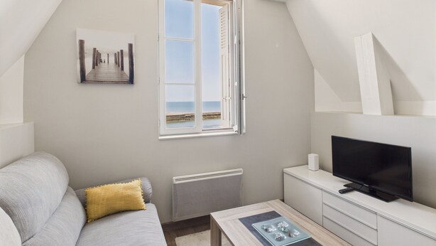 Una habitación moderna y bien iluminada con un ventanal grande, un televisor de pantalla plana, un sofá y una mesita.