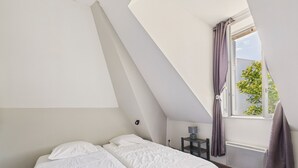 Un dormitorio con techo inclinado, cama con ropa de cama blanca, mesita de noche con lámpara y ventana con cortinas.