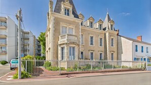 Un edificio histórico con una torre destacada y numerosas ventanas, rodeado por una valla blanca y vegetación.