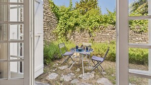 Puerta abierta que da a un patio con pared de piedra, una mesa con sillas y vegetación.