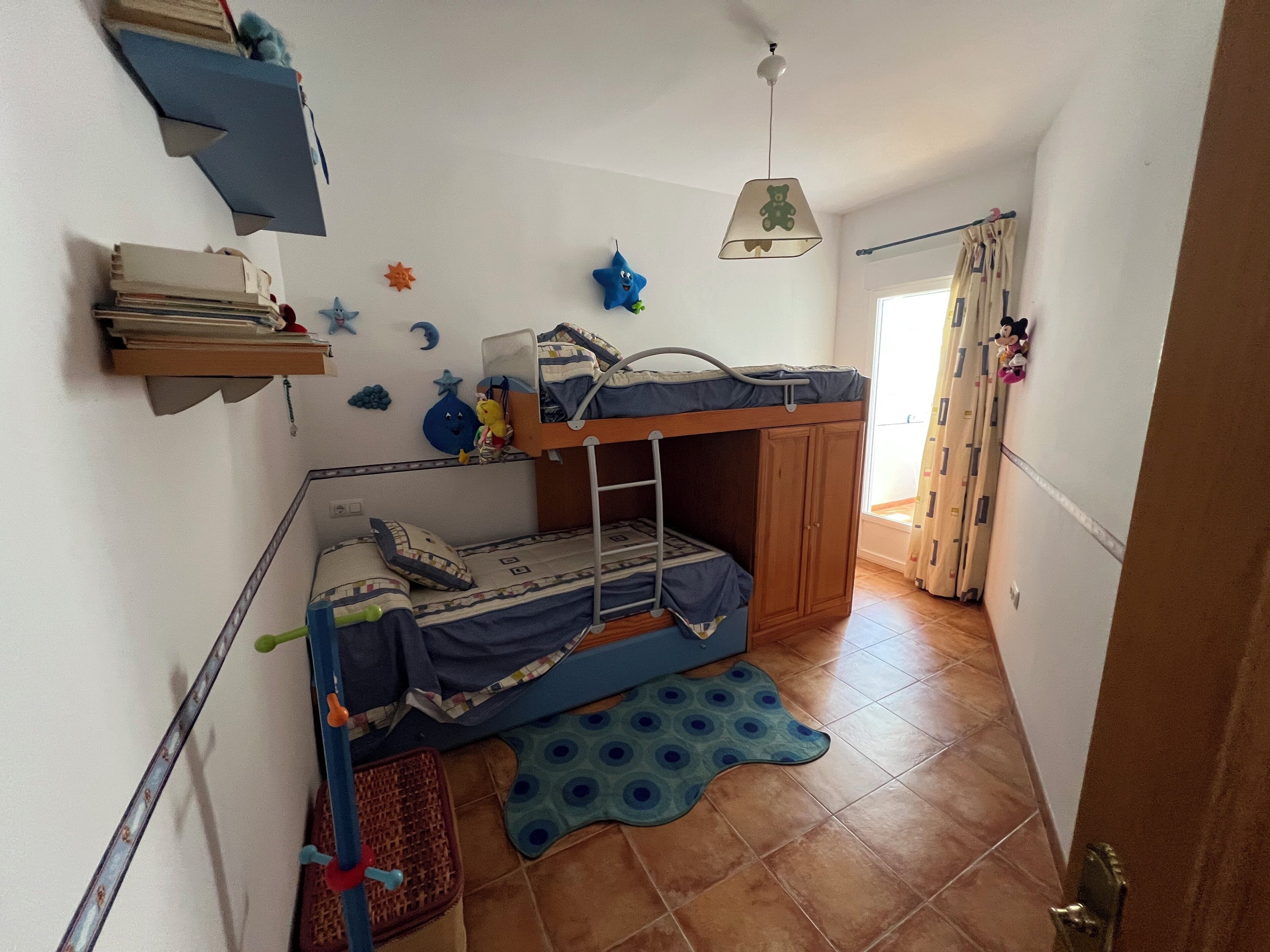 Een slaapkamer met stapelbed, een ladder, een klein plankje met boeken, een ster decoratie en een raam met gordijnen.