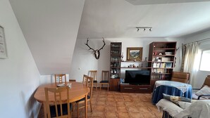 A dining area with a round table and chairs, a television, a bookshelf, and a bed with a plaid blanket. - Magnificent village house surrounded by nature. (Luque)