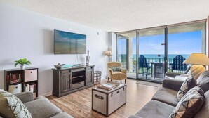 Amplia sala de estar con un gran ventanal que ofrece vistas a la playa, un rincón de descanso acogedor con un sofá y sillones, un mueble de madera y una estantería.