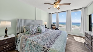 Un dormitorio con cama, mesitas de noche, ventilador de techo, cómoda y vista al mar.