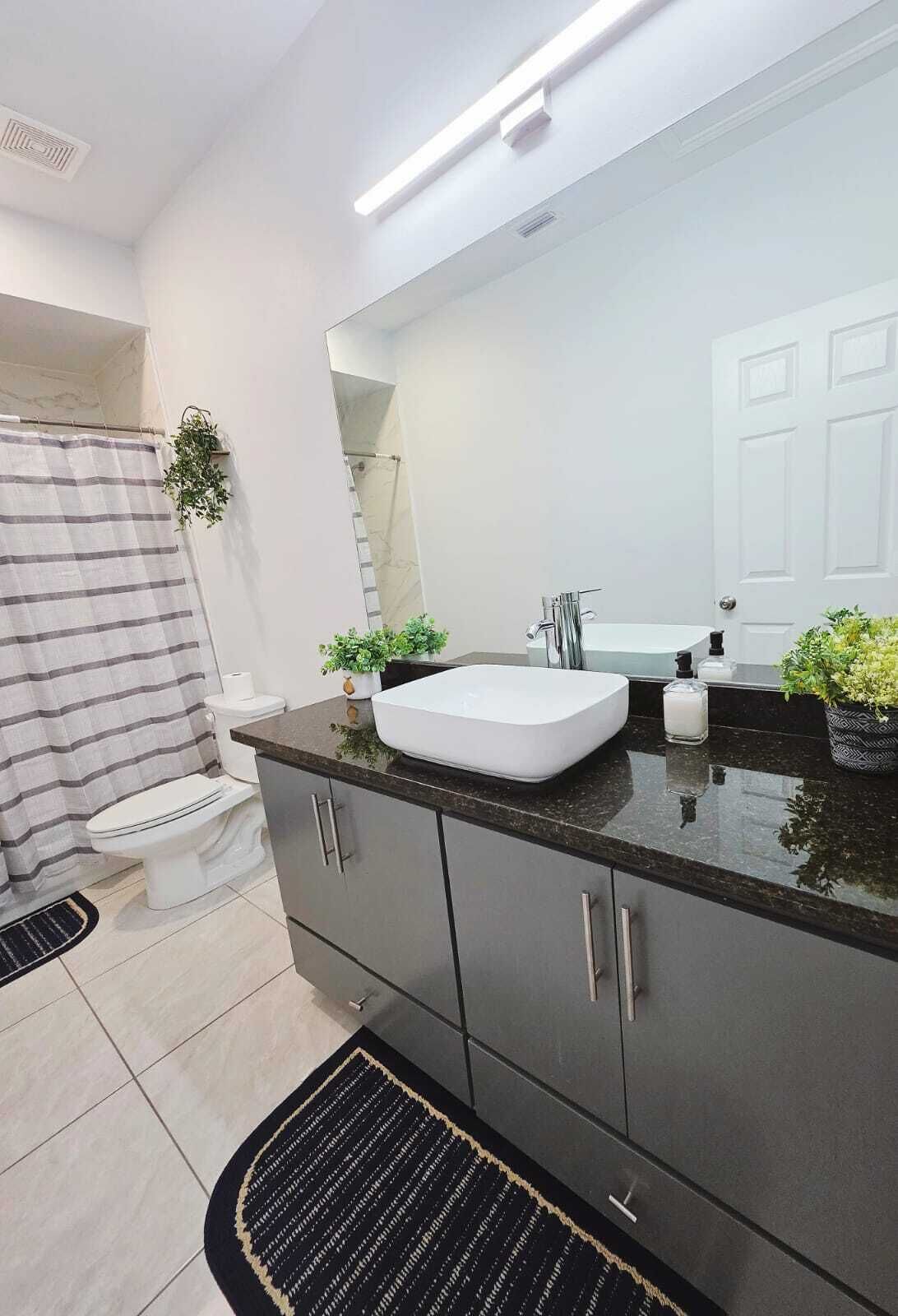 A modern bathroom with a white sink, dark cabinets, a large mirror, and a shower area.