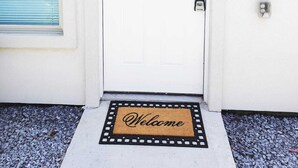 A white door with a 'D' marking, a doormat with 'Welcome' text, a blue window with blinds, and a wall-mounted light fixture.