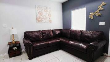 A modern living room with a dark leather sectional sofa, a small side table with a lamp, a decorative wall piece, and a window with blinds.