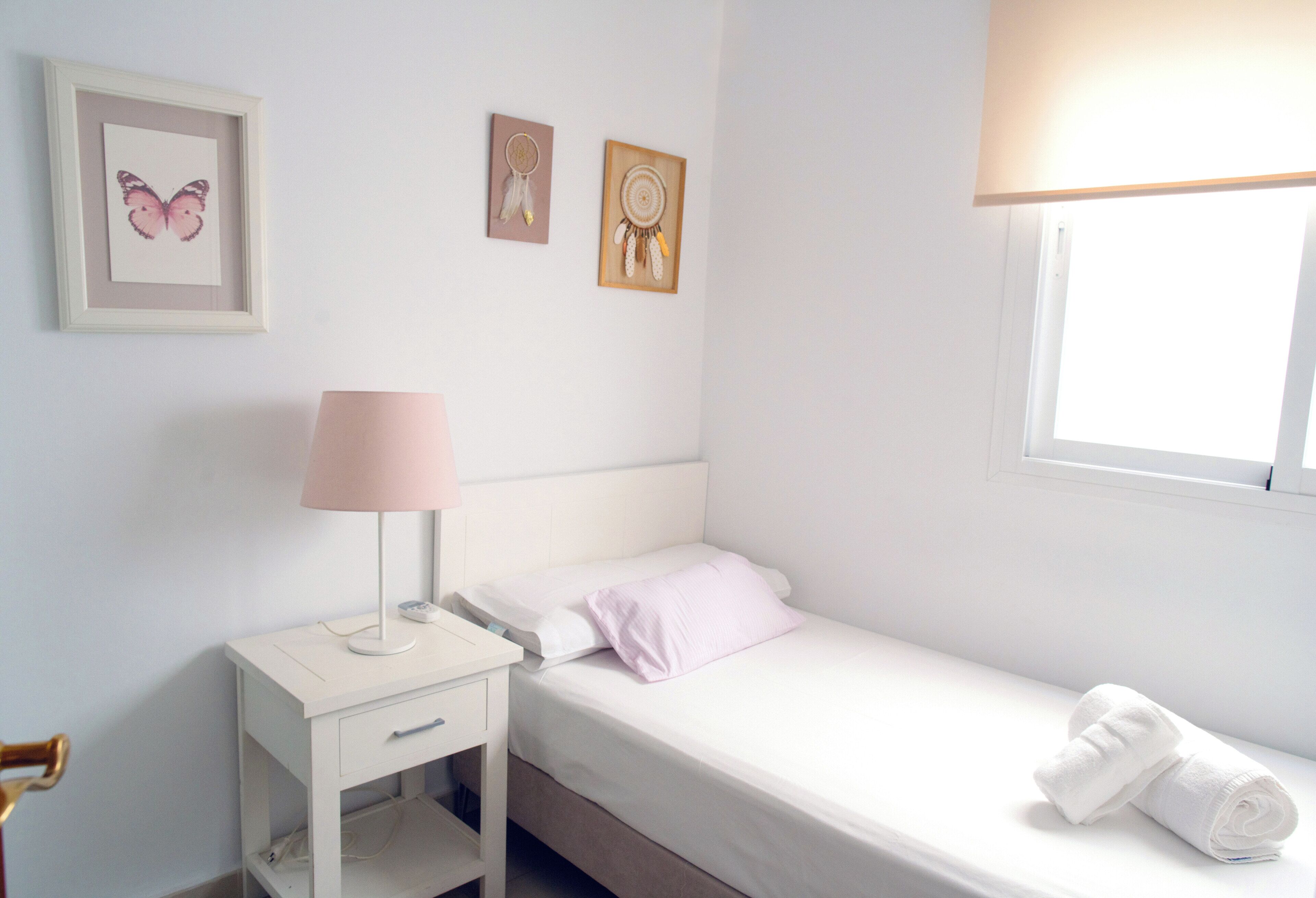 A single-bed room with a bedside table, a lamp, a window, and wall art.