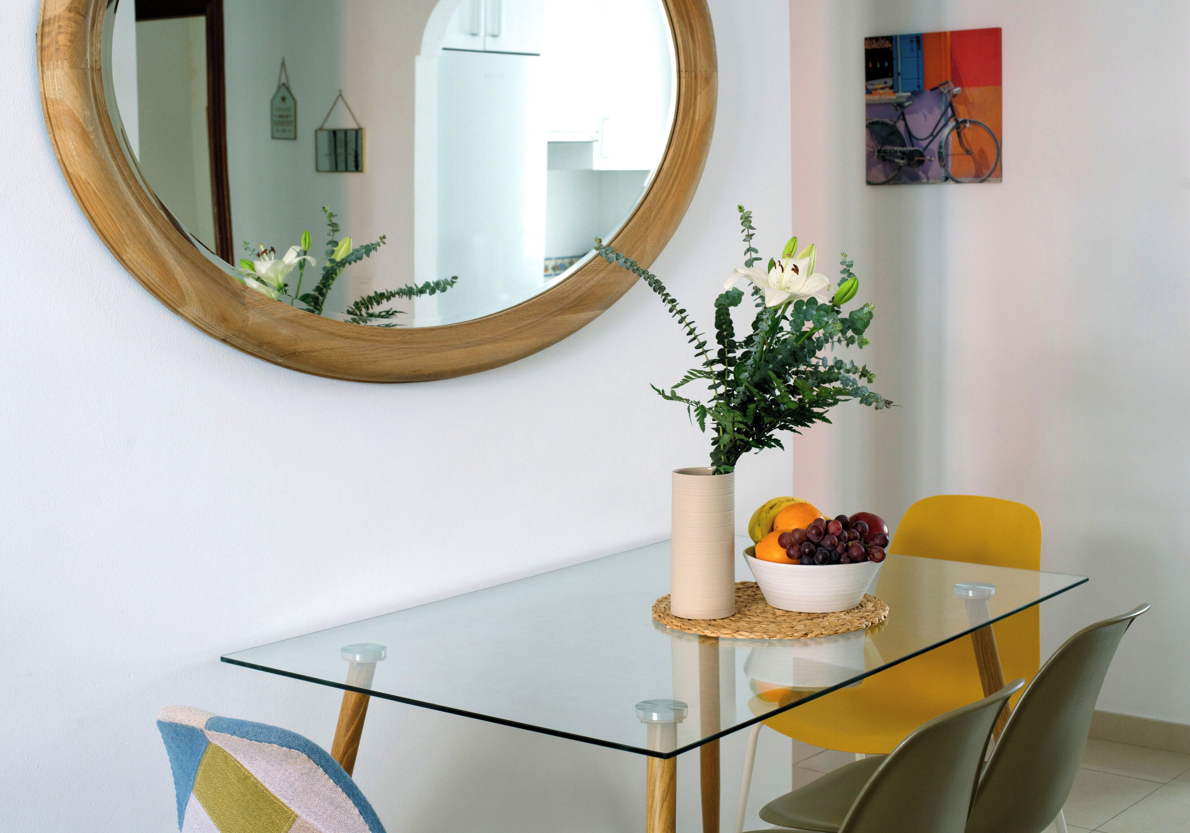Un comedor con una mesa de vidrio, una silla amarilla, un cuenco de fruta y un jarrón con flores.