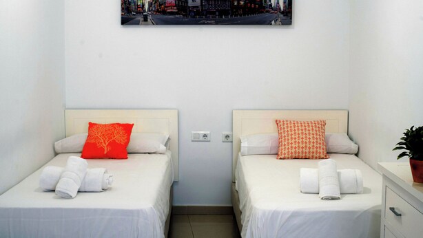 Ett hotellrum med två sängar, båda med vita sängkläder och röda kuddar, en tavla med en stadsbild ovanför sängarna och en krukväxt på nattduksbordet.