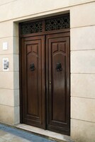 Una puerta doble grande de madera oscura con detalles de metal ornamentado en su parte superior, ubicada en una fachada de edificio color beige.