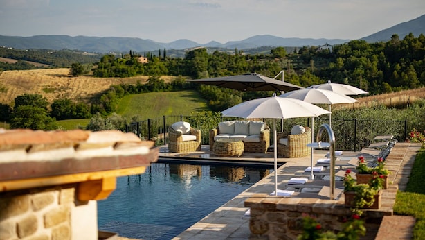 Uma área à beira da piscina com mobiliário de vime, guarda-sóis e vista para o campo.