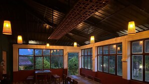 Un comedor con piso a cuadros, mesas y sillas de madera. Se ven grandes ventanales con vistas a follaje y un edificio.