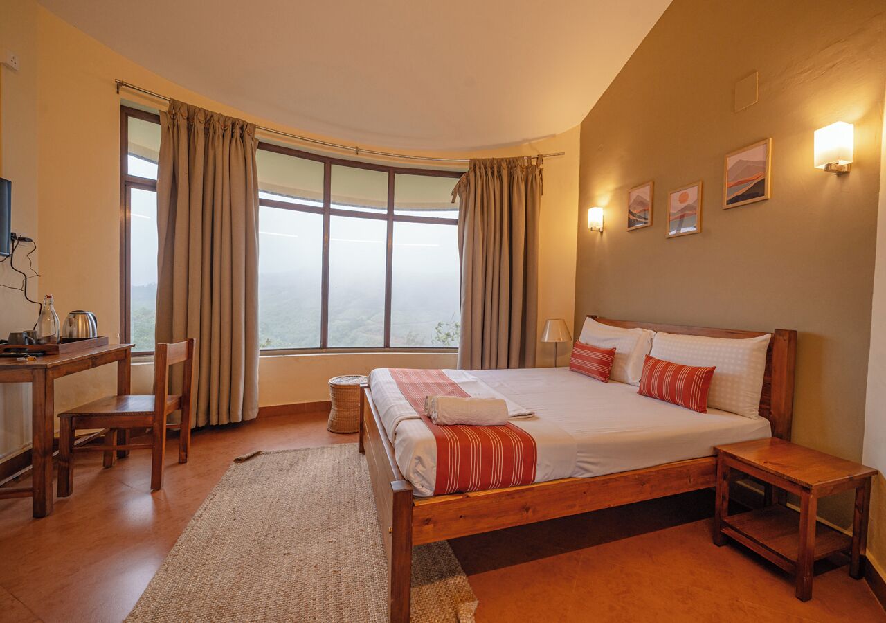 A bedroom with a large bed, a desk, and a chair. There is a window with curtains, a lamp, and wall art.