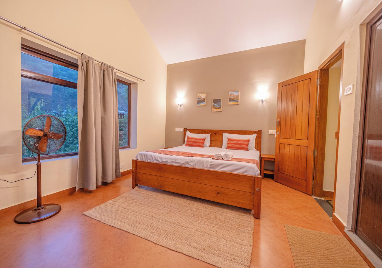A bedroom with a wooden bed, a standing fan, a window with curtains, and a wooden wardrobe.