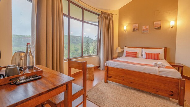 A bedroom with a wooden bed, a desk with a remote control, a lamp, and a view of the outdoors. - Aruvi Vagamon by Voye Homes (Vagamon)