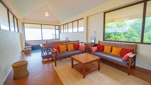 A spacious living area with a large window, a wooden sofa set, a coffee table, and a wicker basket. - Aruvi Vagamon by Voye Homes (Vagamon)