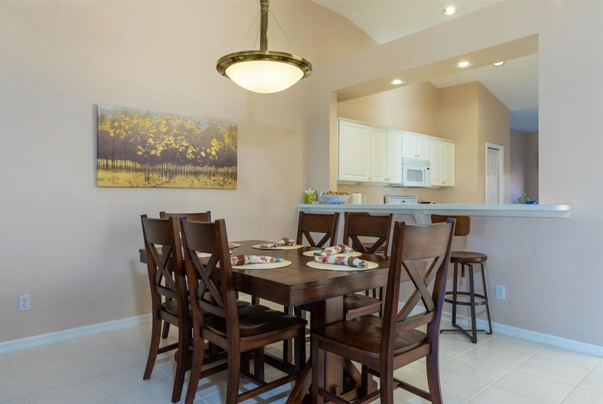 A dining area with a wooden table and chairs, a painting on the wall, and a kitchen in the background.