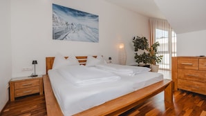 A modern bedroom with a wooden bed, white linens, a nightstand, a lamp, a large window with curtains, a potted plant, and a painting of a snowy mountain scene. - Wonderful private villa for 10 guests with WIFI, TV, terrace and pets allowed (Zell Am See)