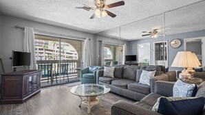 Un salón amplio con un sofá grande, una mesa de centro de vidrio y una televisión. Hay un balcón con vistas a edificios y palmeras.
