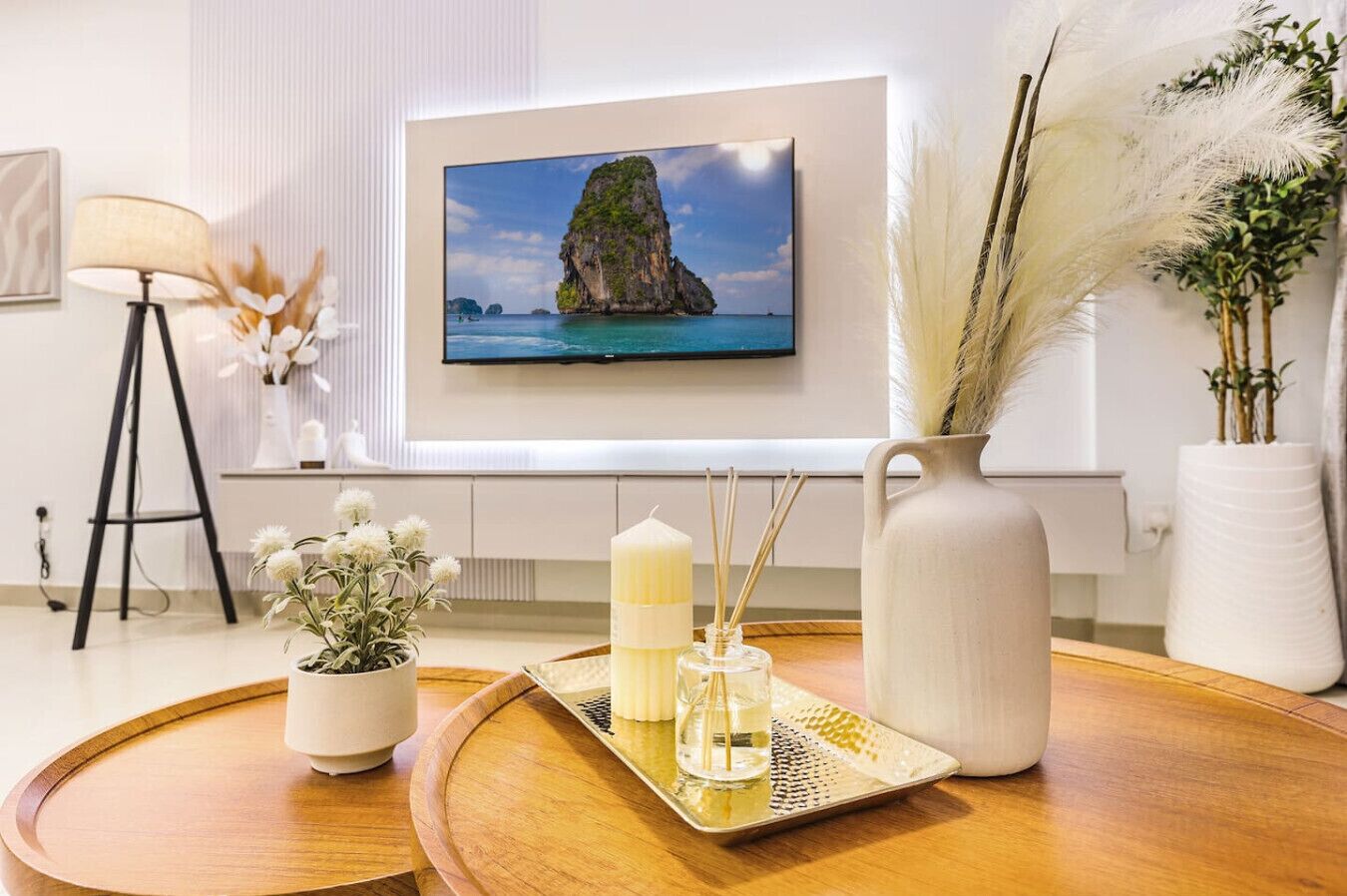 Une chambre moderne avec une grande fenêtre, un tableau encadré représentant un paysage côtier, un lampadaire, une plante en pot, un plateau avec des bougies et des diffuseurs à roseaux, et un vase contenant des plumes d’herbe des pampas.