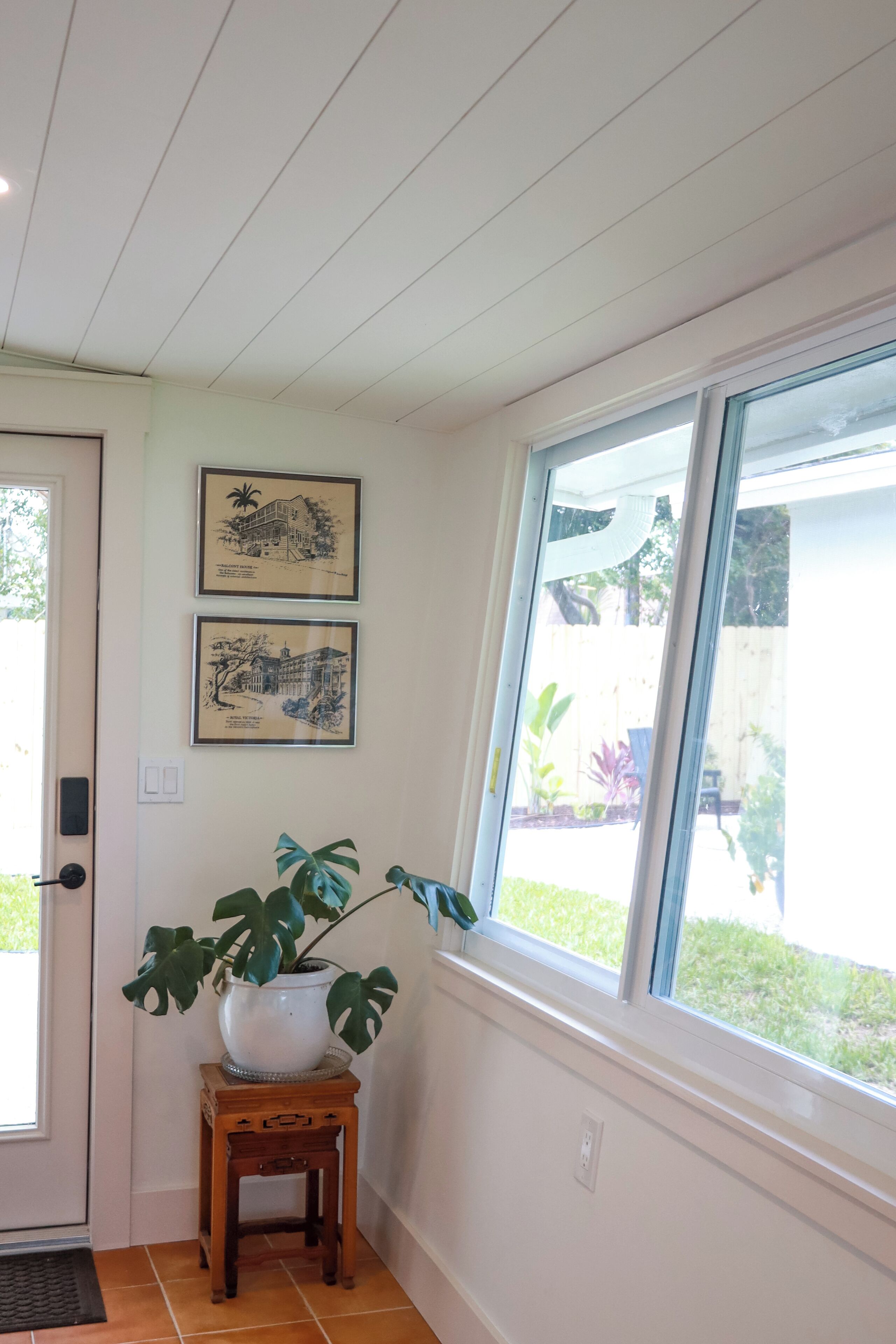 A room with a wooden door, a window with a view of a garden, and two framed pictures on the wall.