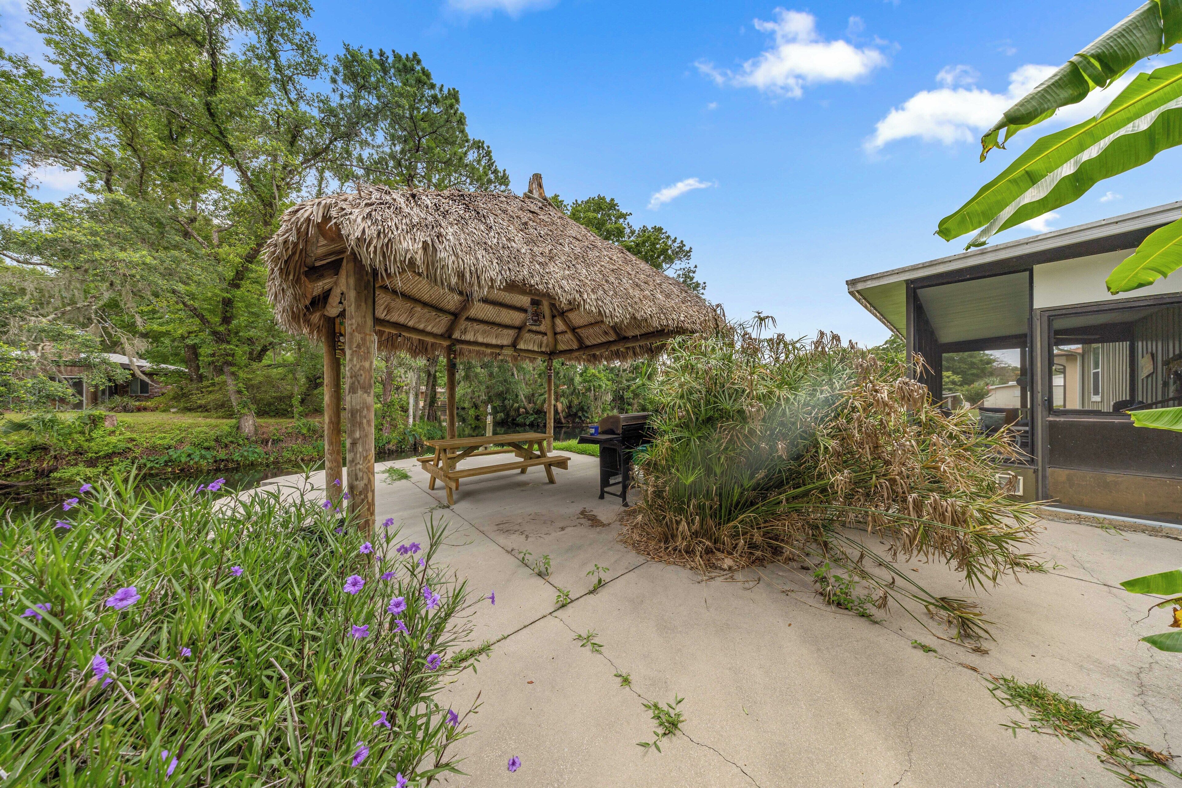 Ein überdachter Außenbereich mit einer Holzbank, einem Reetdachbau und einem Garten mit verschiedenen Pflanzen.