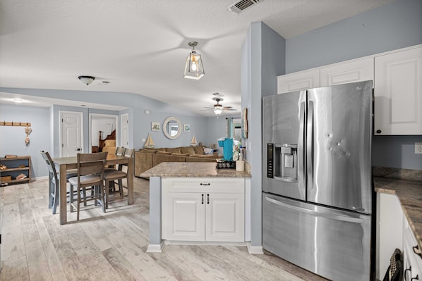 A kitchen with white cabinets, a stainless steel refrigerator, a dining area with a wooden table and chairs, and a ceiling fan.