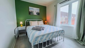 A neatly made bed with striped bedding, two bedside lamps, a small table, and a window with curtains.