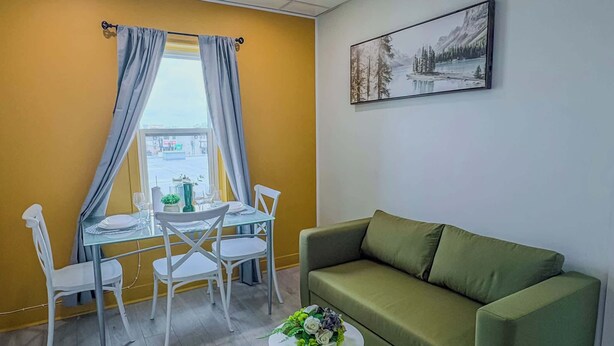 A room with a green sofa, a dining table with chairs, a small round table with a flower arrangement, and a painting on the wall.