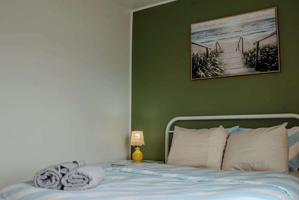 Una cama bien hecha con ropa de cama a rayas, una toalla enrollada y una lámpara de noche sobre una mesita.