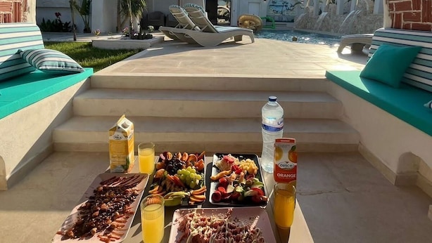 A poolside breakfast setup with various fruits, meats, and beverages, overlooking a modern building with arches and a clear sky. - Dar maryem (Hammam Sousse)