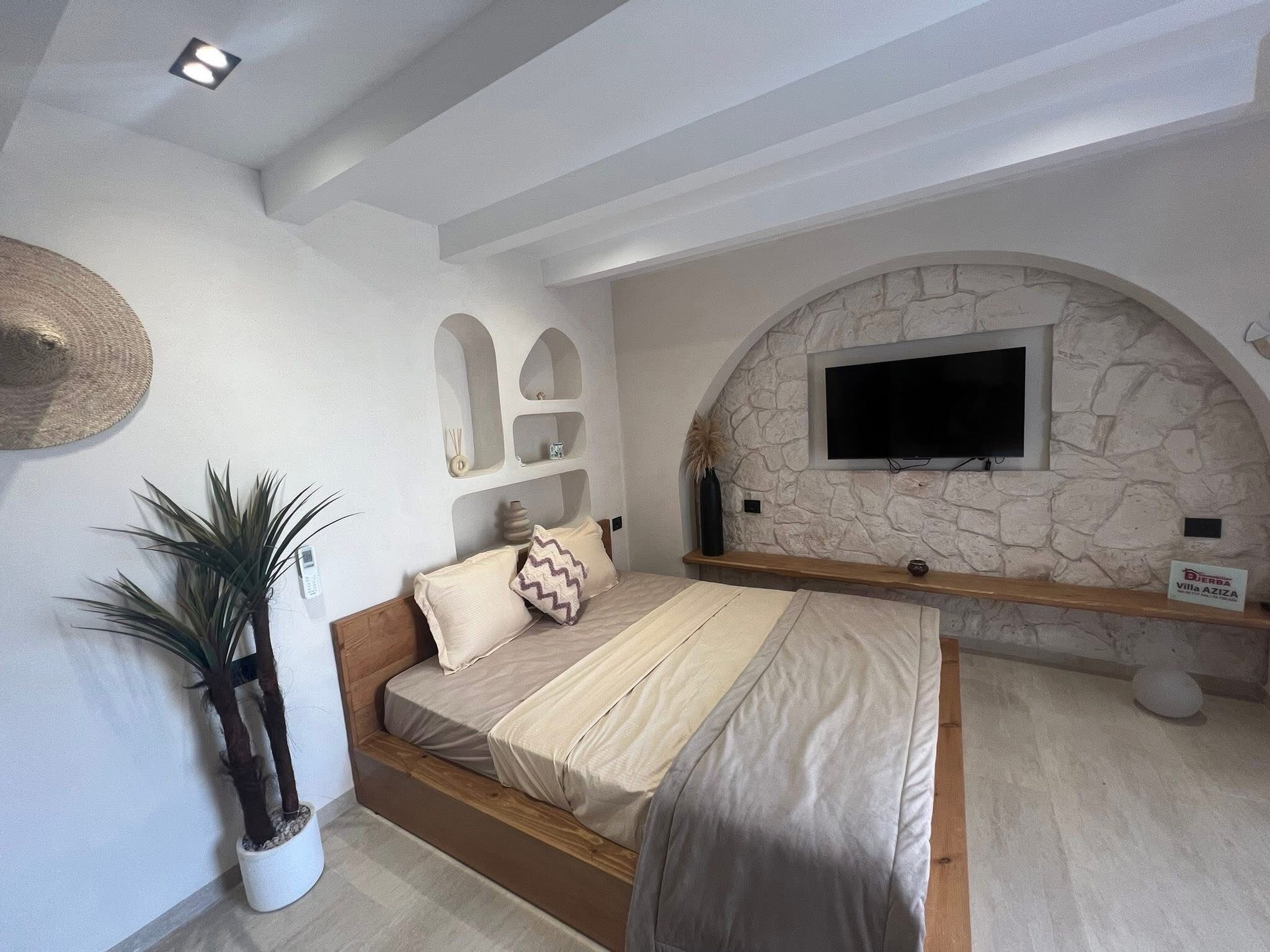 Un dormitorio con pared de piedra, un televisor montado sobre un estante de madera, una cama con ropa de cama color beige y una planta en maceta.