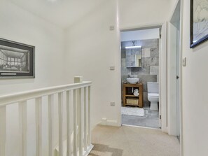 Un couloir avec une rambarde blanche, un tableau encadré au mur et une porte donnant sur une salle de bain avec des toilettes blanches et un mur en pierre.