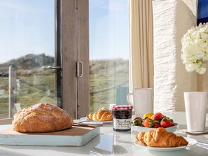Une table dressée pour le petit-déjeuner avec des croissants, des fraises, un pot de confiture et une vue sur un paysage verdoyant par la fenêtre.